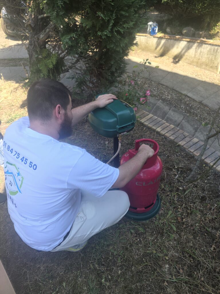 Technicien IJN+ raccordant la bouteille de propane d'un piège Mosquito Magnet installé sous un arbre, dans un jardin de Conflans-Sainte-Honorine