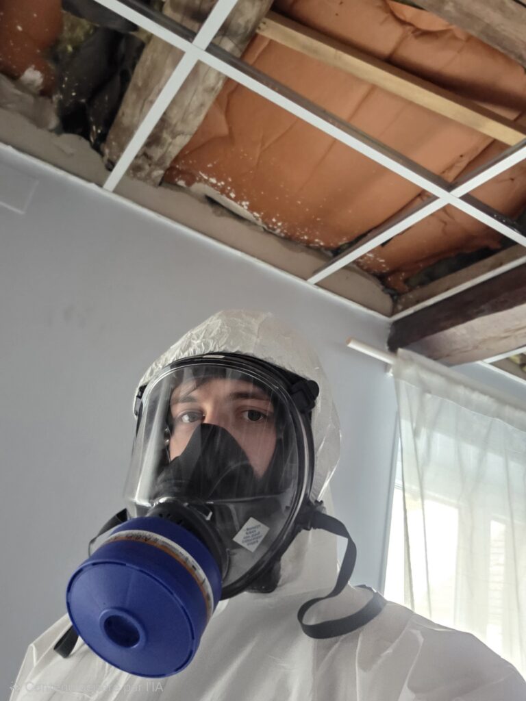 Technicien IJN+ en combinaison et masque respiratoire à cartouche sous une charpente en bois, lors d'un traitement contre les vrillettes à Limay